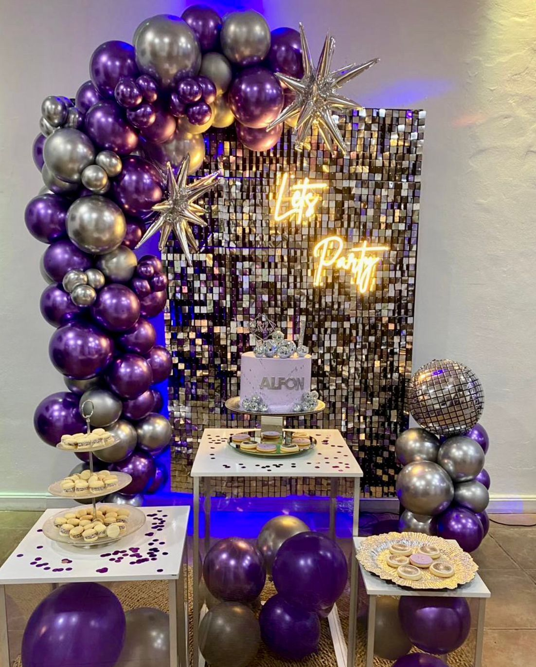 Exhibición de postres para fiestas en color púrpura y plateado con globos, letrero de neón y una mesa para pasteles.