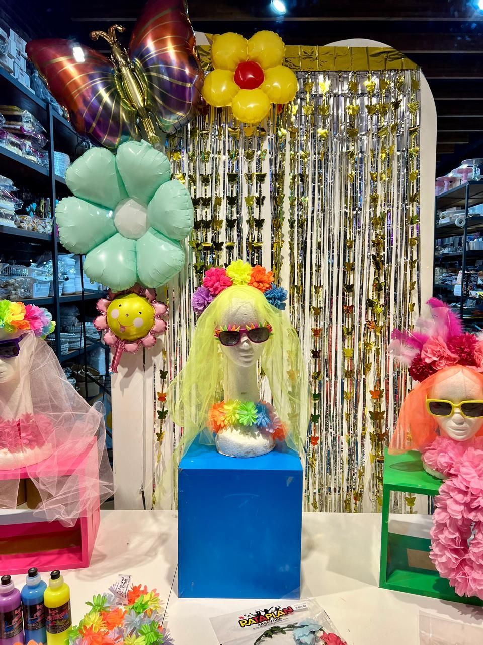 Exhibición de pelucas y gafas de sol coloridas con decoraciones de globos y fondo de lentejuelas.