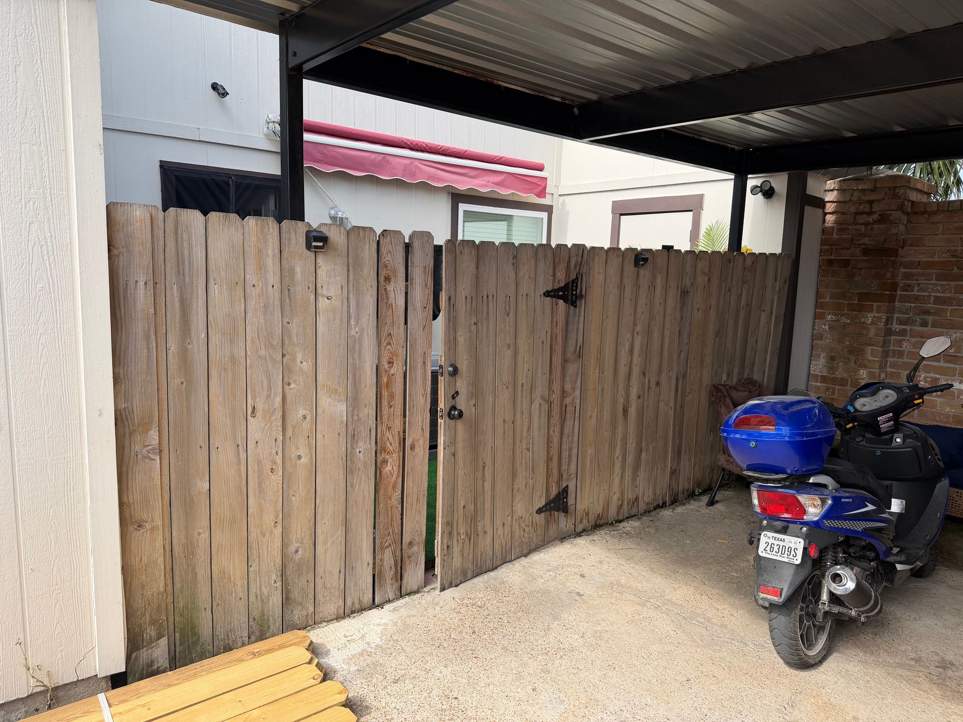 Wooden gate under a carport beside a parked blue motorcycle on gravel.