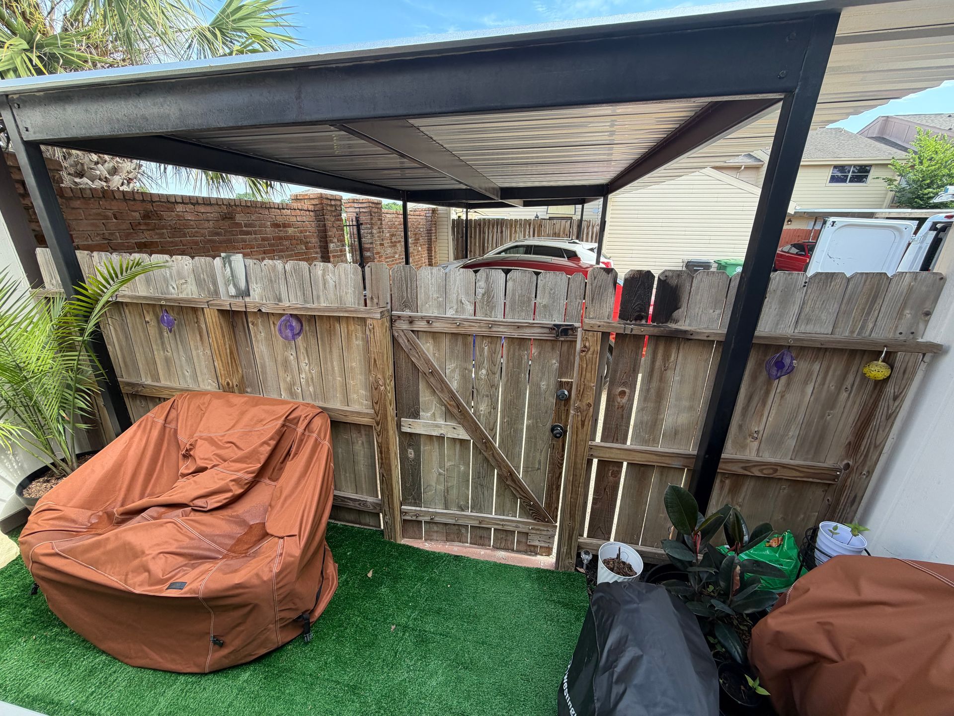 Backyard with a wooden gate, metal canopy, artificial grass, and covered patio furniture.