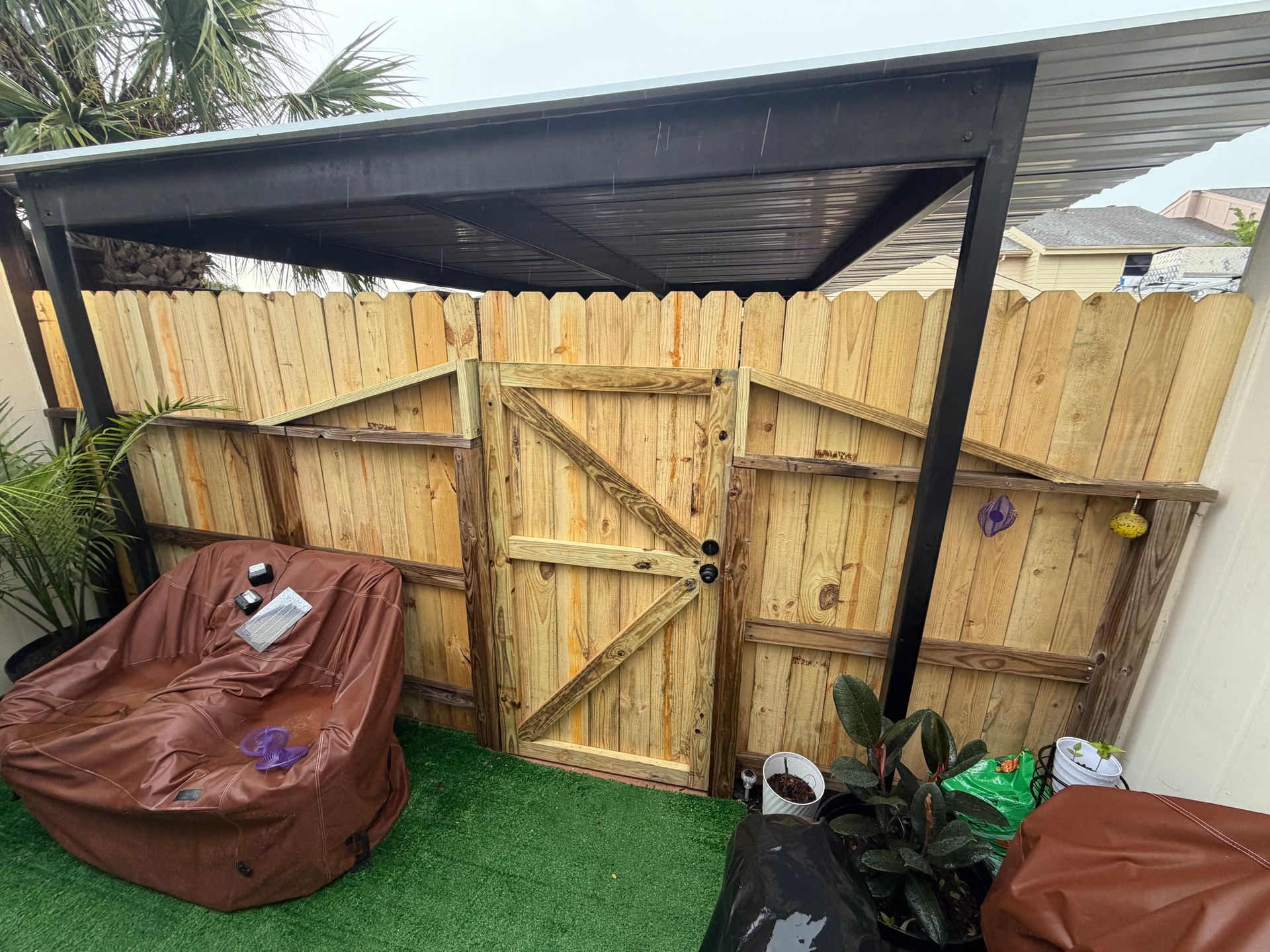Backyard wooden gate under a black canopy, with covered items on artificial grass on both sides.