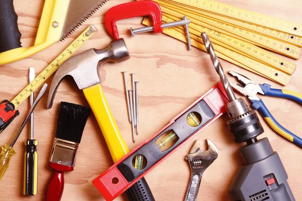 Assorted tools on a wooden surface, including hammer, level, saw, drill, tape measure, and brushes.