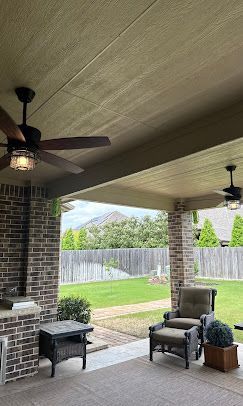 Covered backyard patio with brick grill, ceiling fan, chairs, and a grassy fenced yard beyond.