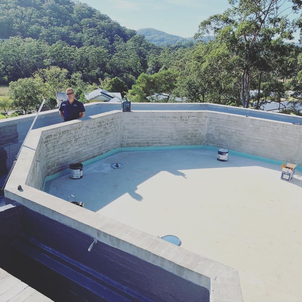 A Man Is Standing On The Roof Of A Building Overlooking A Swimming Pool — Coastwide Waterproofing in Woy Woy, NSW