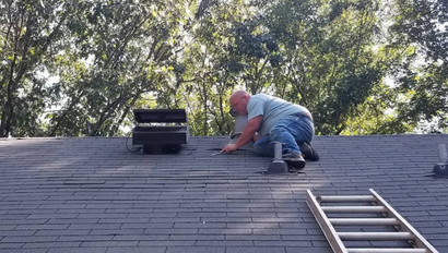 Worker kneeling on a shingled roof, repairing near a vent with a ladder below.
