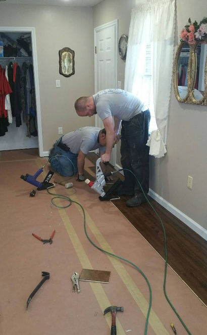 Two workers installing flooring in a hallway with tools and extension cords.