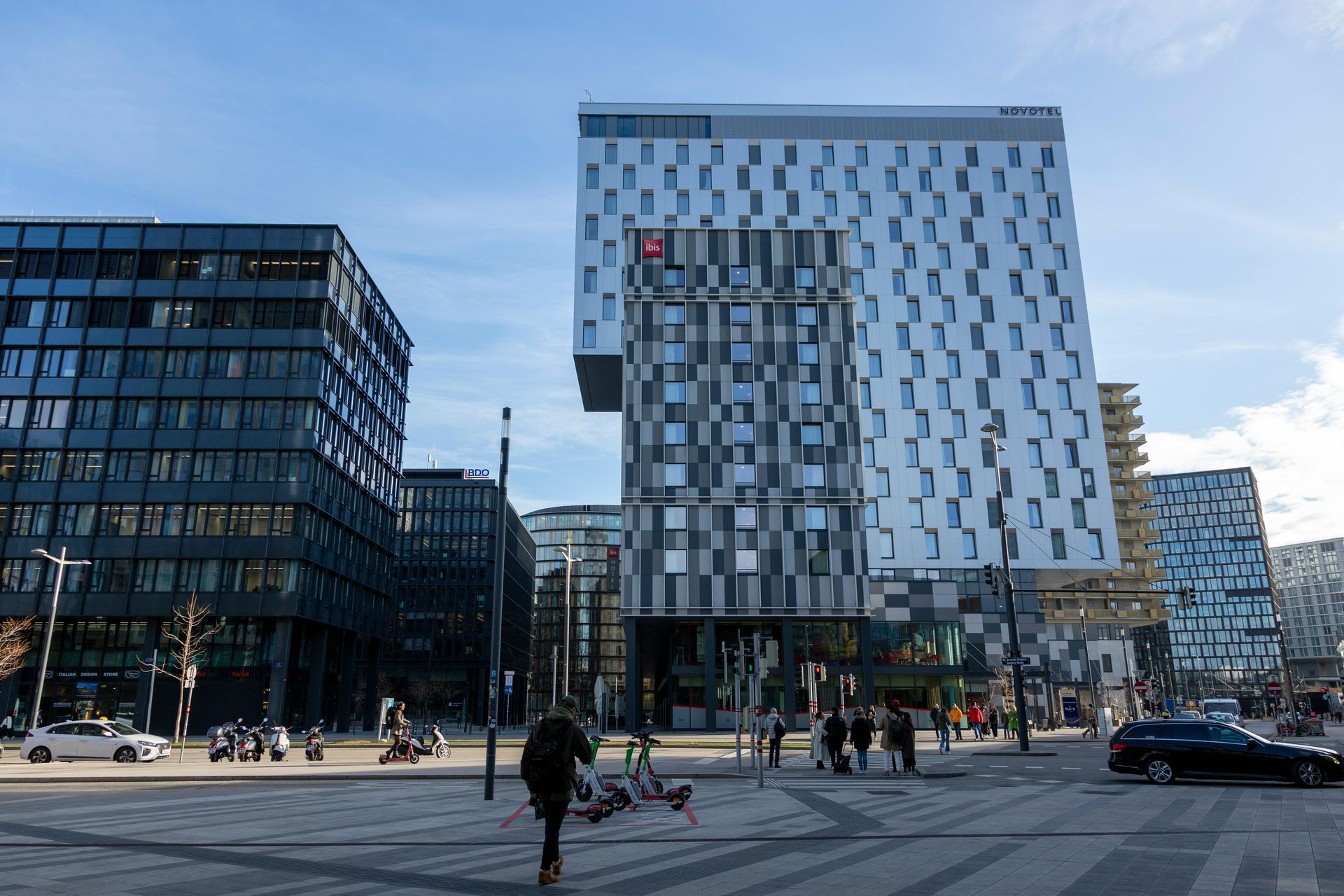 Modern glass office buildings in a city plaza with a few pedestrians and blue sky