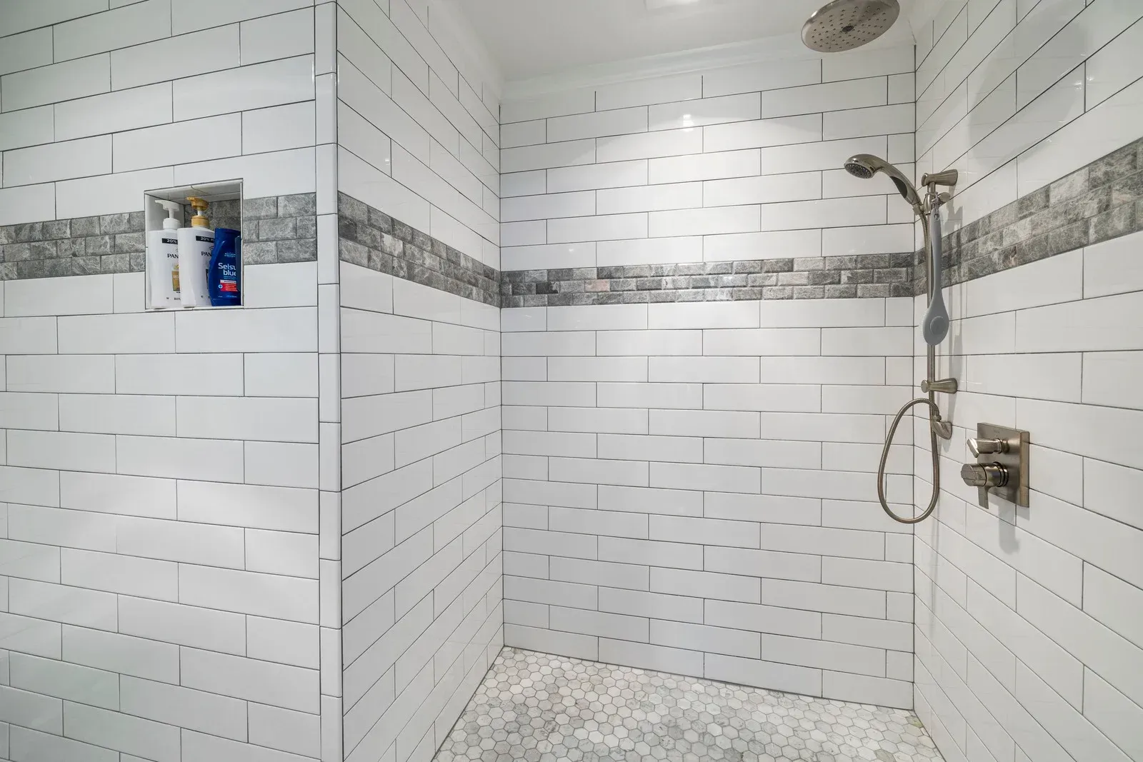 White-tiled shower with gray stone accent band and recessed shelf holding shampoo and soap.