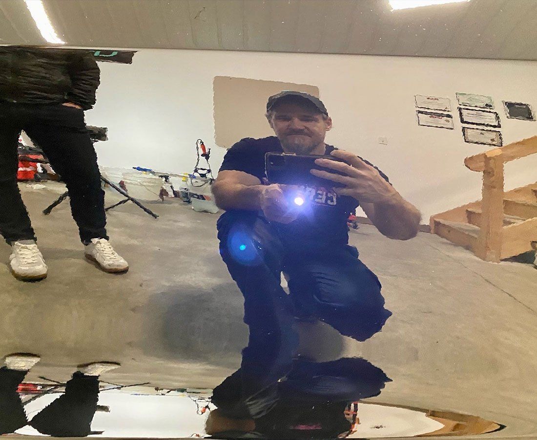 Man crouching to take a mirror selfie in a room, wearing a cap and dark clothes, with bright camera flash.