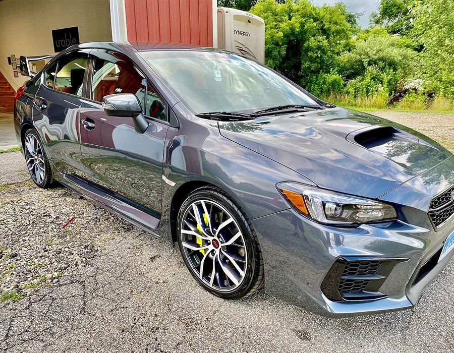Gray sports sedan parked on gravel beside a red building and greenery