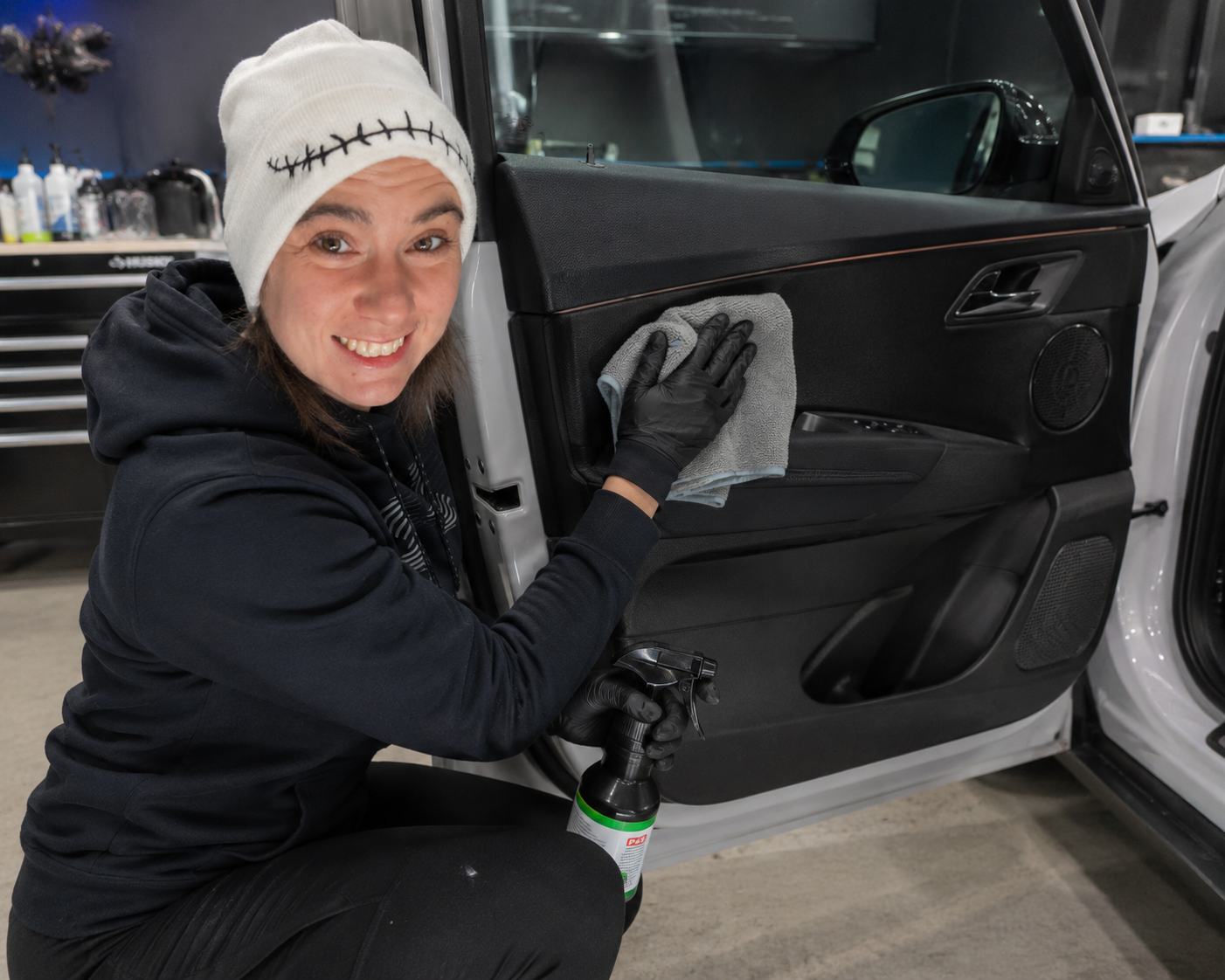 Woman in a black hoodie and white beanie cleaning a car door interior with a cloth in a garage.