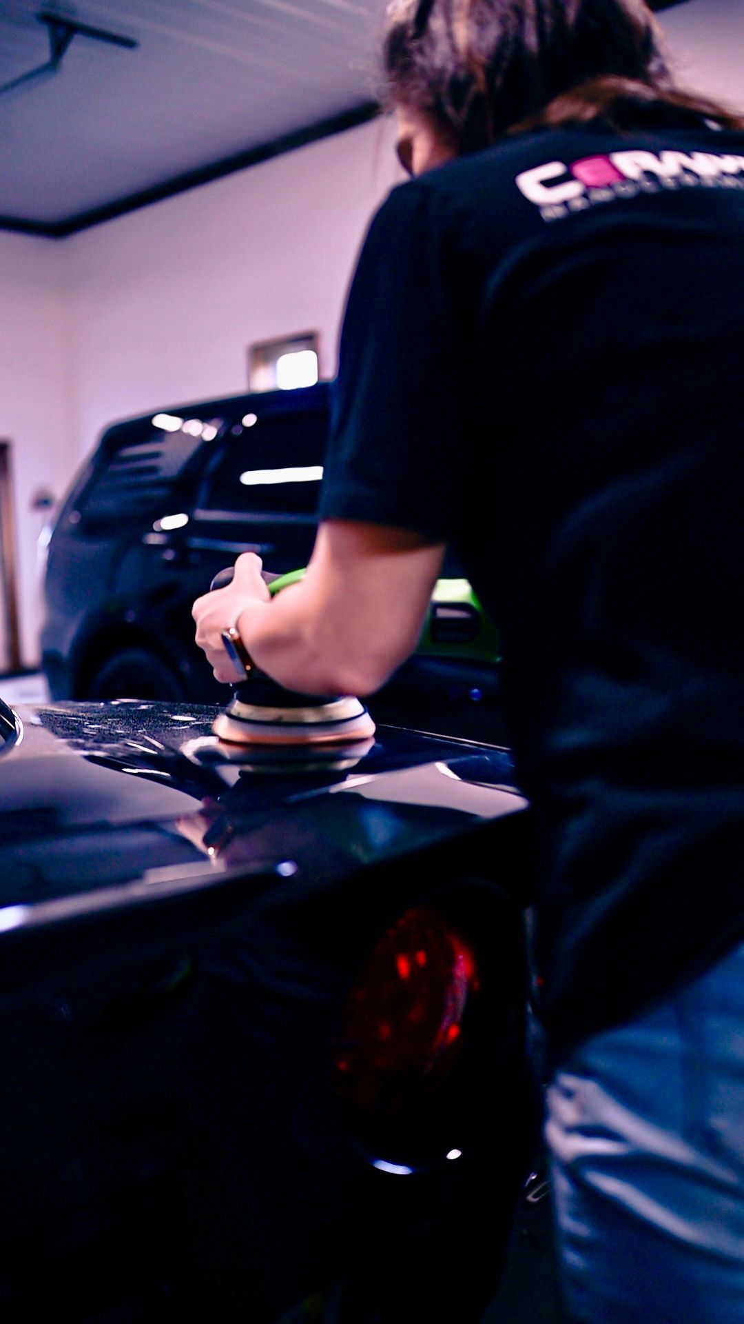 Person polishing a black car in a garage, wearing a black shirt and blue jeans.