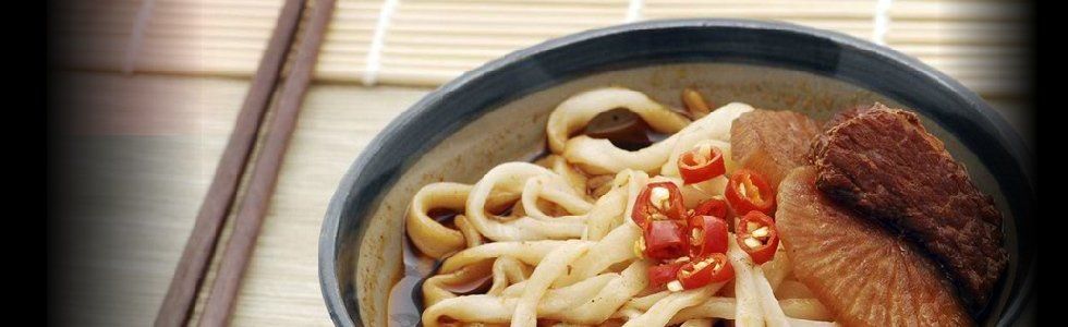 Una ciotola di spaghetti con carne a fette e guarnizione rossa su un tavolo di legno.