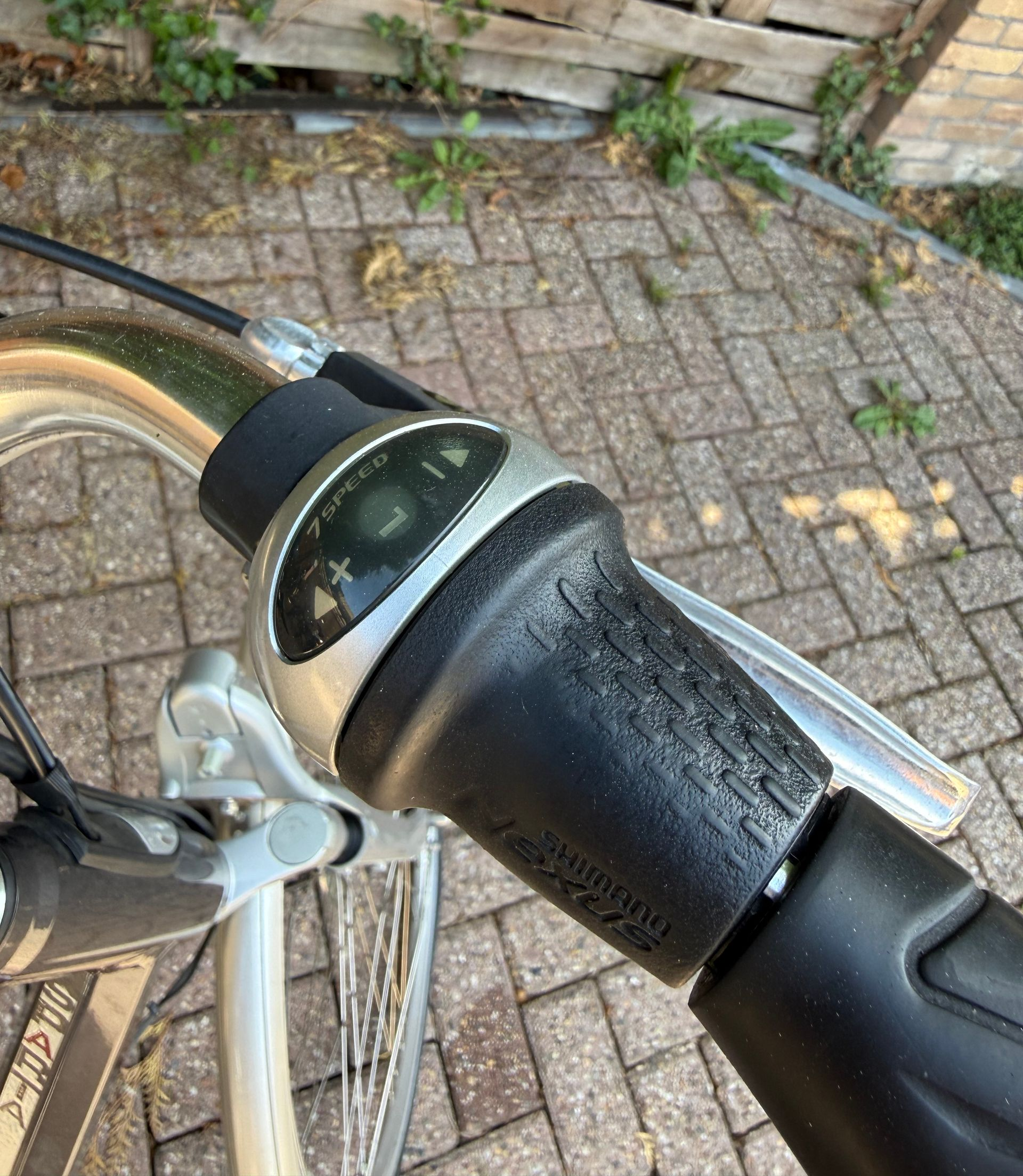 Close-up van een fietsstuur met een zwarte, gestructureerde handgreep en versnellingspook op een straat met kinderkopjes.