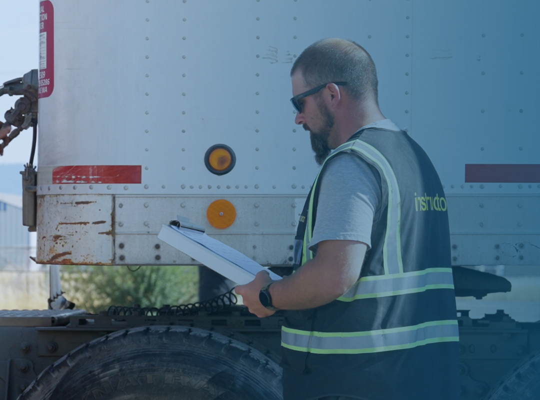 A CDL instructor in a safety vest is standing next to a truck, holding a clipboard and instructing.