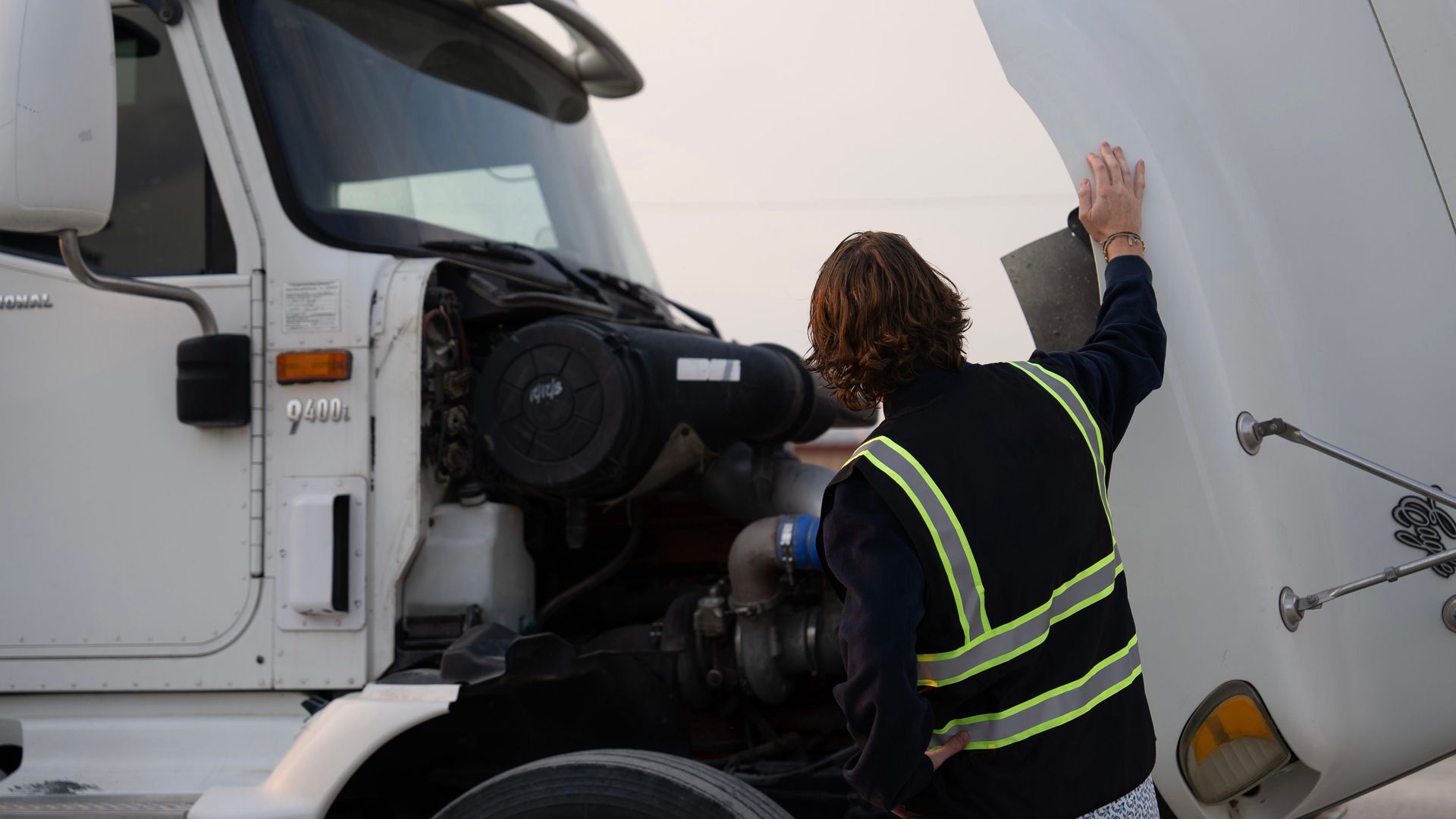 Two cdl students in high-visibility vests work on the landing gear of a semi-truck trailer outdoors.