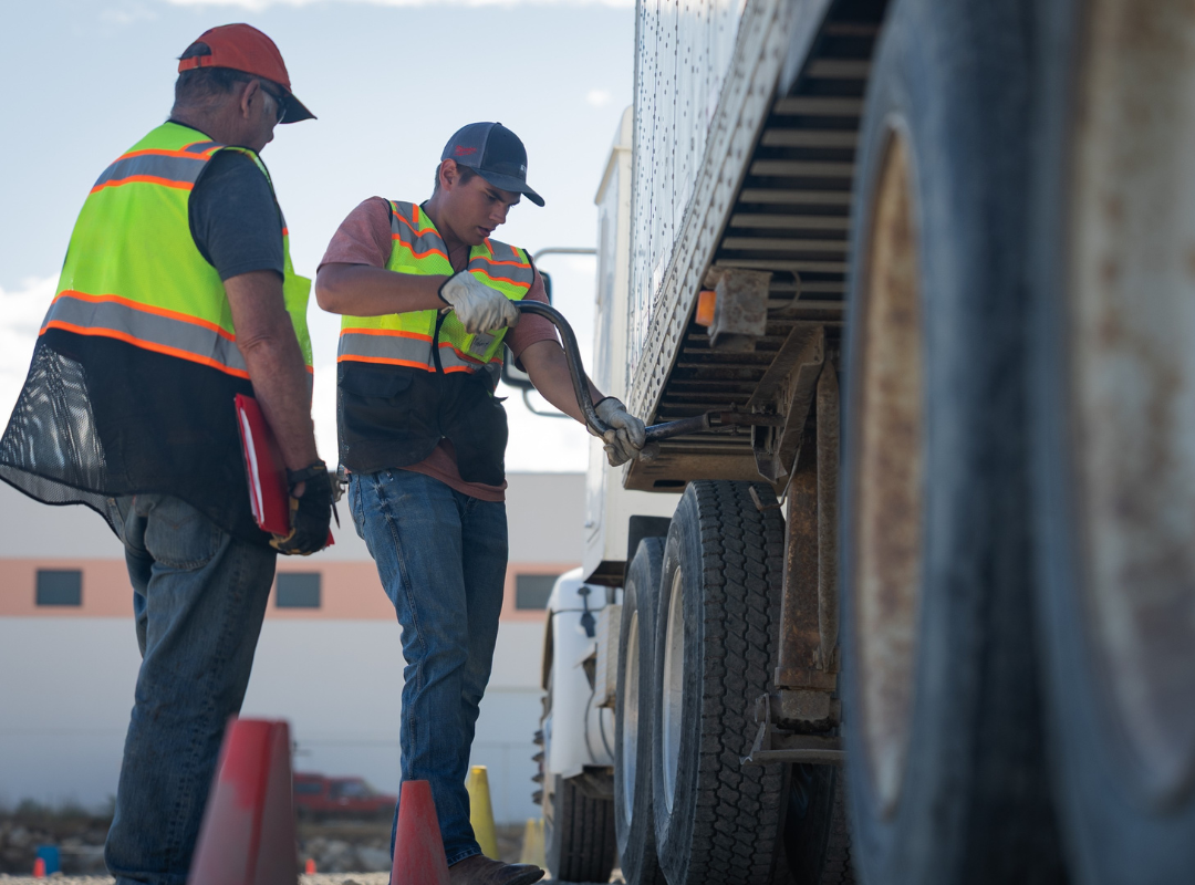 Two cdl students in high-visibility vests work on the landing gear of a semi-truck trailer outdoors.