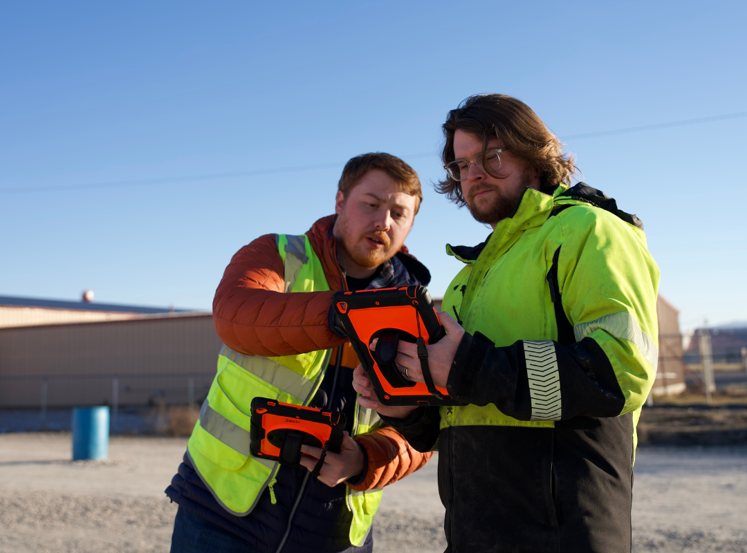 Two workers in high-visibility jackets holding equipment outdoors under a clear blue sky