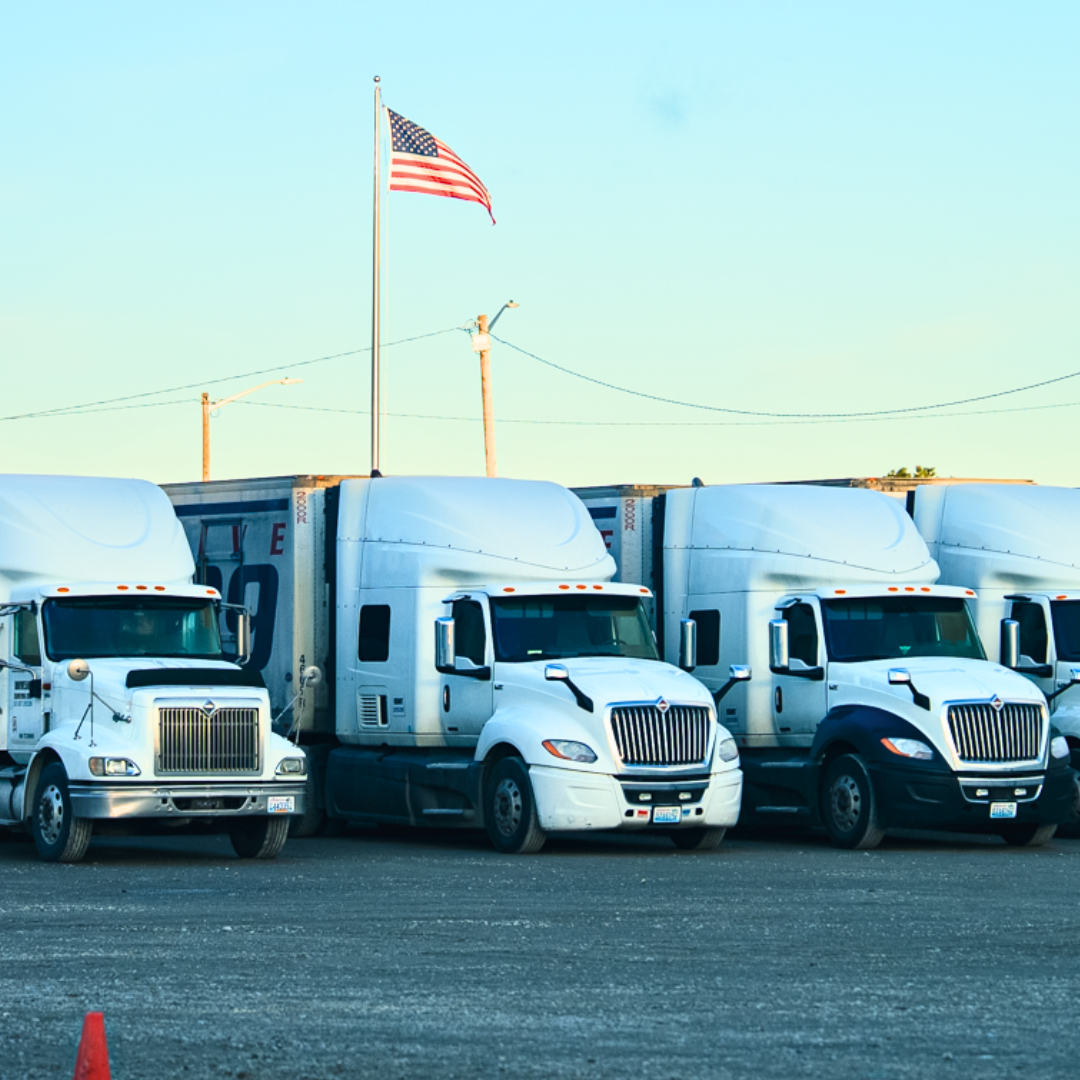White semi-trucks parked in a lot under a blue sky, American flag waving.