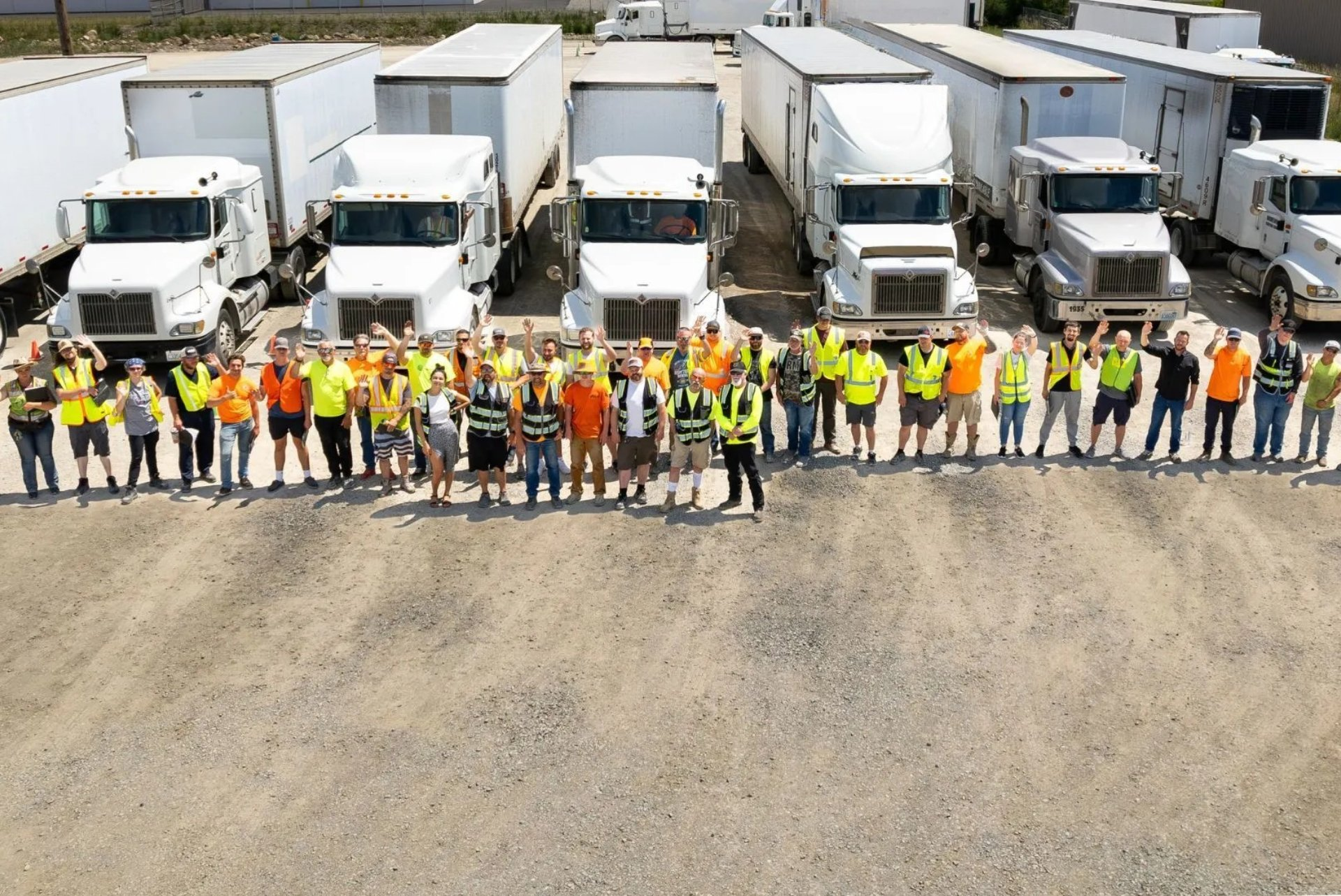 Aerial view of CDL students and instructors in safety vests standing in front of a fleet of training semi trucks