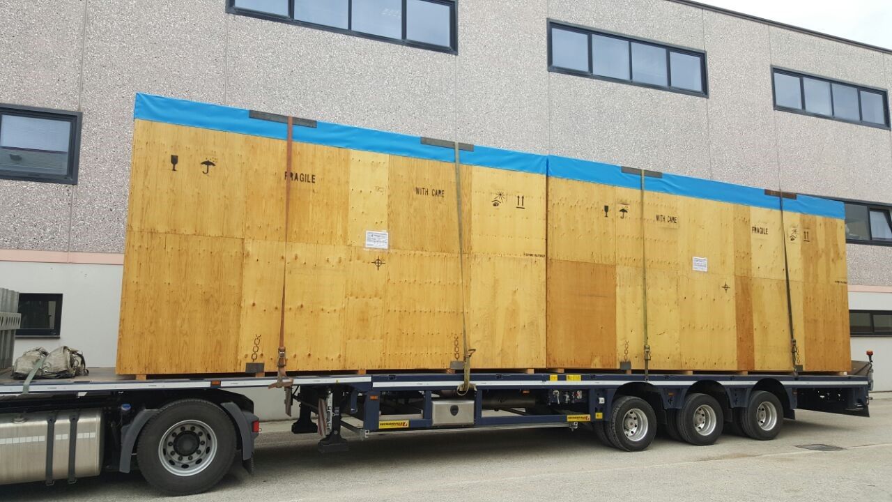 Una grande cassa di legno gialla su un rimorchio a pianale, parcheggiata accanto a un edificio grigio.