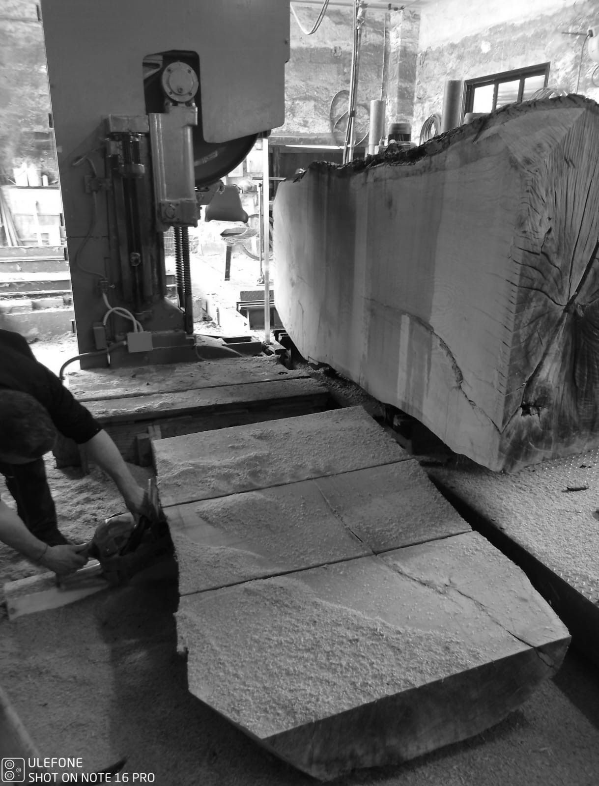 In un'officina, una grande sega industriale taglia una lastra di pietra in blocchi, con polvere e macchinari ben visibili.