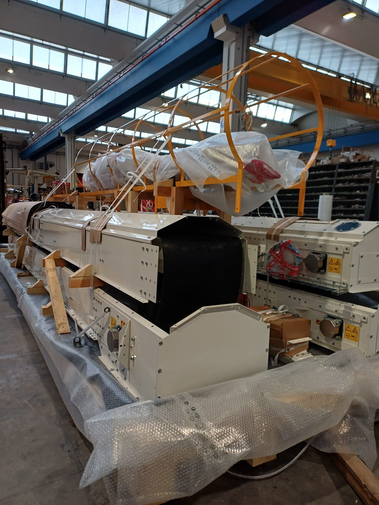 Macchinari industriali avvolti in plastica e schiuma all'interno di un magazzino o di una fabbrica, sospesi tramite attrezzature aeree.