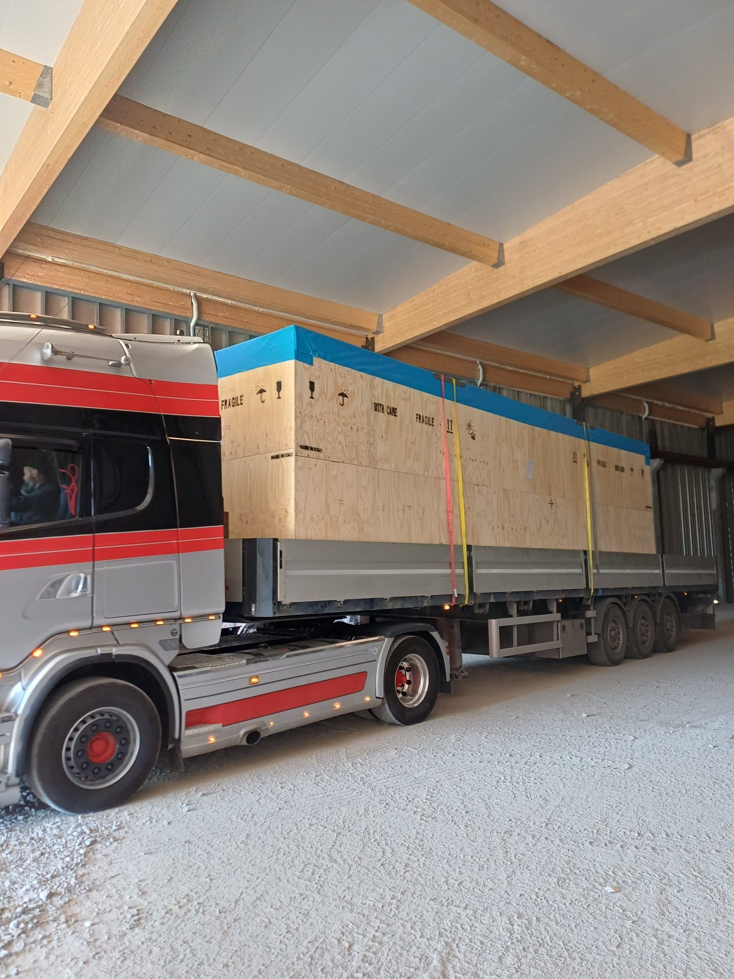 Un camion semirimorchio rosso e grigio trasporta una grande cassa di carico avvolta all'interno di un magazzino con tetto in legno.