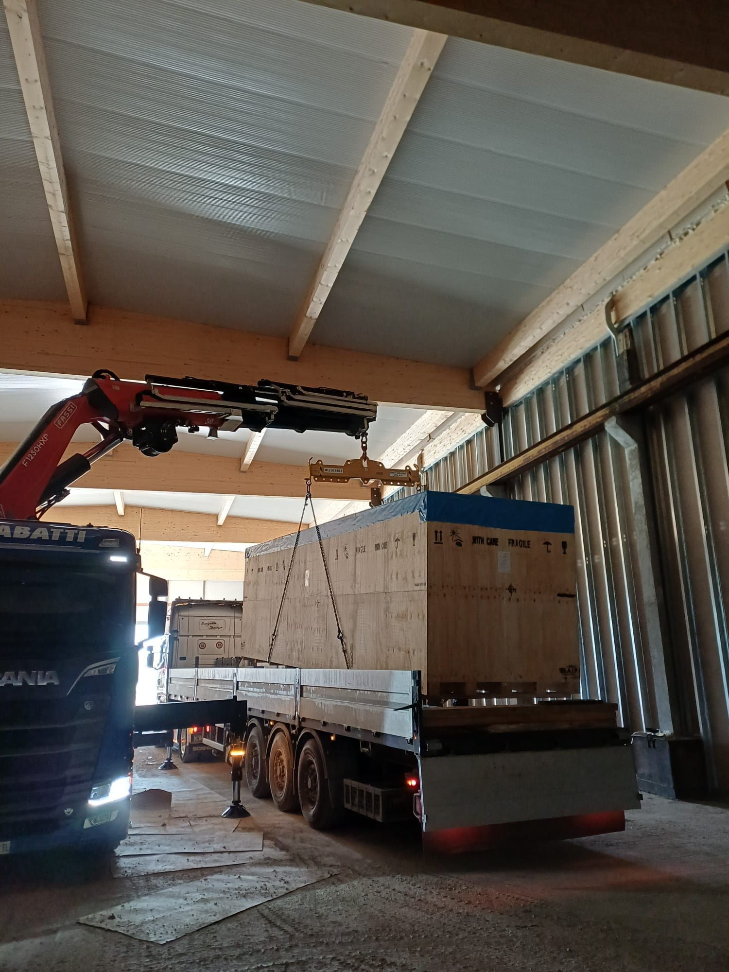 Una gru carica un camion a pianale con una grande cassa di legno all'interno di un magazzino.