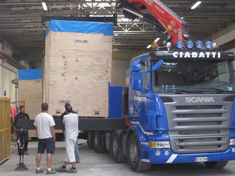Un camion Scania blu carica una grande cassa di legno in un magazzino mentre due persone guardano
