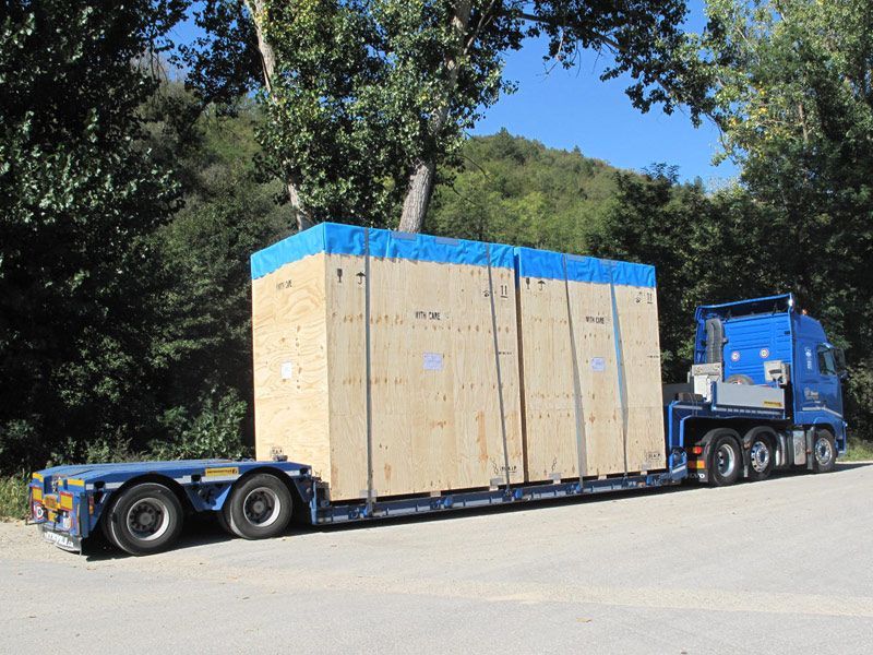 Un camion semirimorchio blu trasporta una grande cassa di legno su un rimorchio a pianale lungo il ciglio della strada.