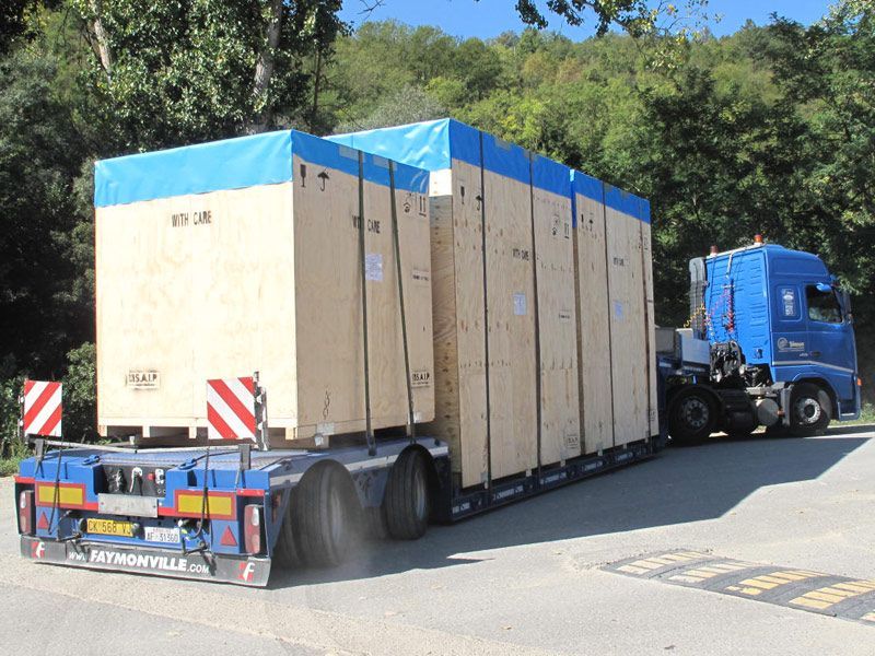 Un camion blu con semirimorchio che traina un alto rimorchio beige per il trasporto merci su una strada fiancheggiata da alberi e colline.
