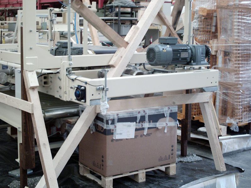 Macchina industriale con telaio metallico e motore che solleva una scatola di cartone sul pavimento di una fabbrica.