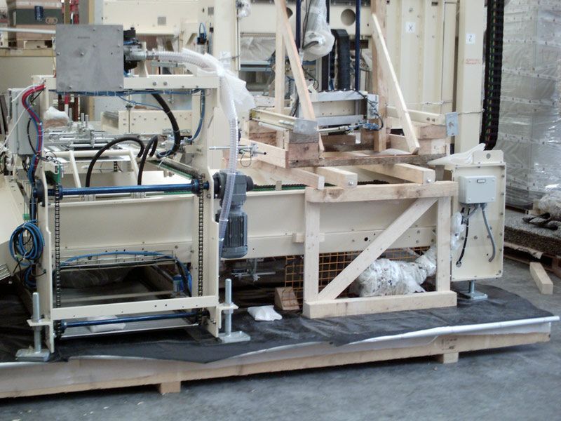 Macchinario industriale in un impianto di produzione, con struttura metallica, tubi flessibili e una piattaforma simile a un nastro trasportatore.