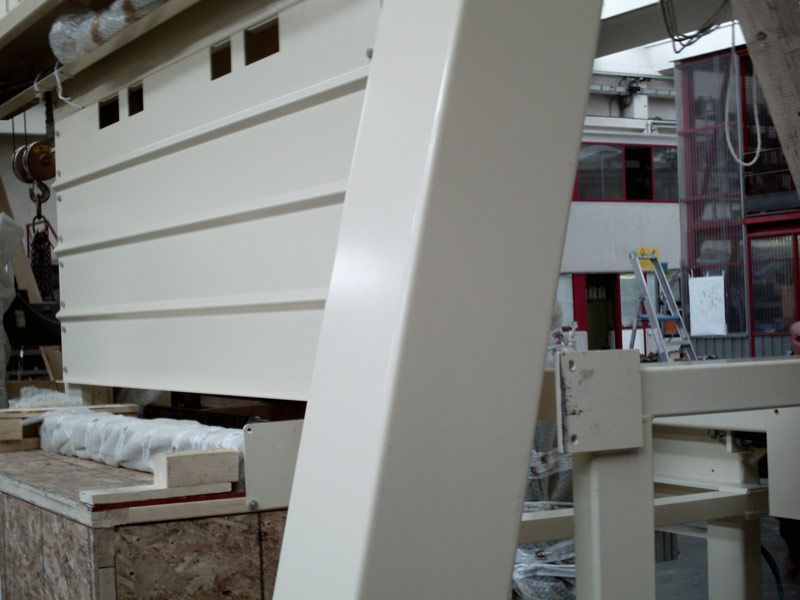 In un'officina, un grande pannello industriale bianco viene sollevato, con macchinari e impalcature visibili.