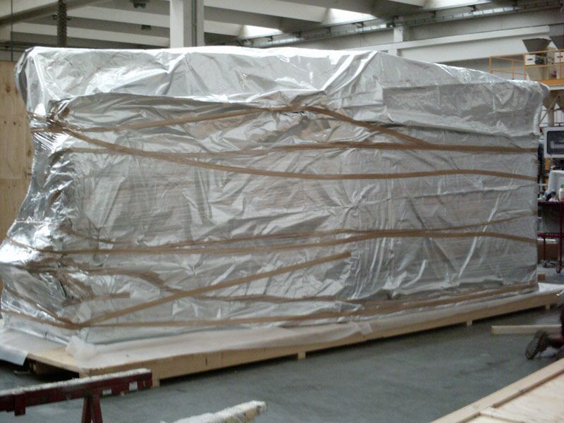 Un grande pallet avvolto in una pellicola, posizionato su una piattaforma di legno in un magazzino, è ricoperto di plastica argentata e legato con cinghie.