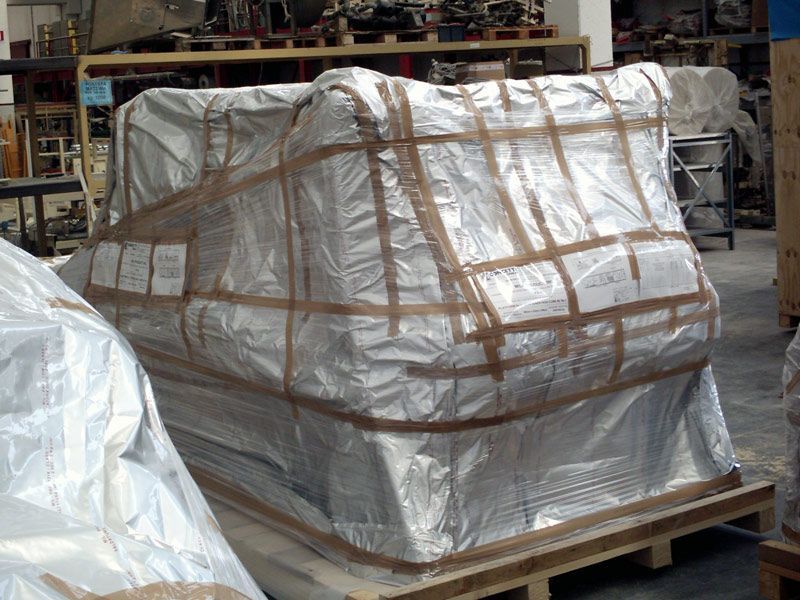 Un grosso oggetto bianco, avvolto in un involucro e pallettizzato, è fissato a un pallet di legno in un magazzino.