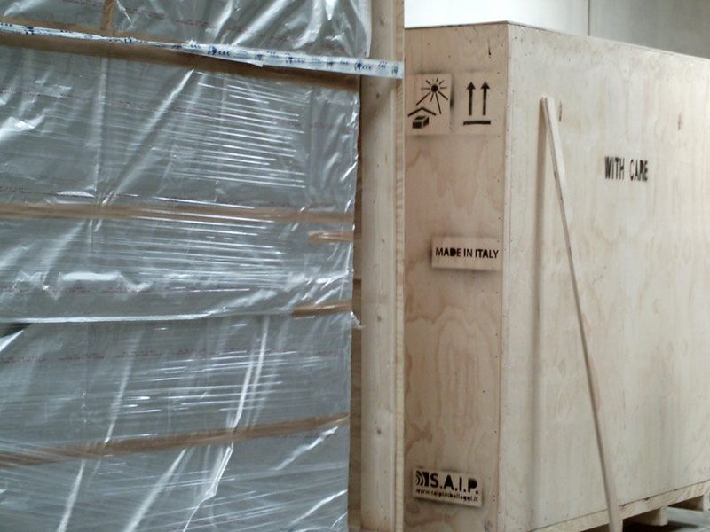 Cassa di legno per spedizioni, posizionata accanto a una parete rivestita di plastica, contrassegnata da simboli e etichette di movimentazione.
