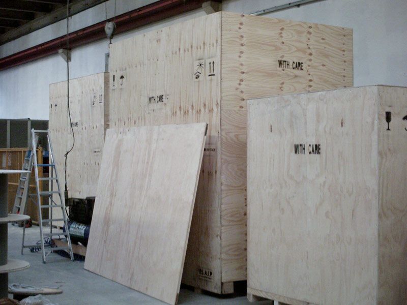 Grandi casse di legno per spedizioni in un magazzino, con una scala e pannelli di compensato nelle vicinanze.