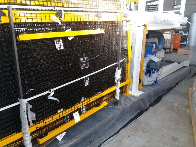 Macchinari industriali con protezioni in rete nera, binari gialli e pavimento grigio in un'area di fabbrica.