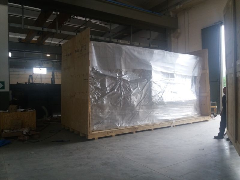 Una grande cassa avvolta in un imballaggio protettivo si trova su un pallet in un magazzino poco illuminato, con una persona in piedi vicino all'ingresso.
