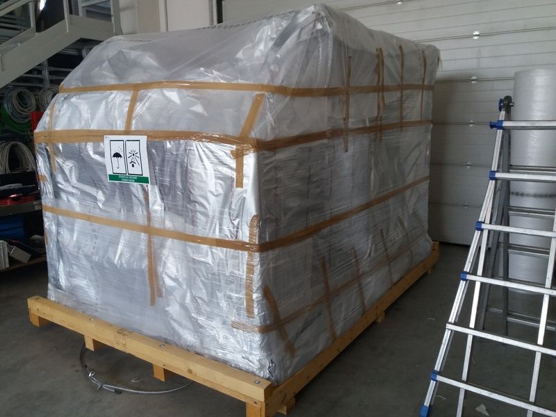 Un grande pallet avvolto in un telo protettivo si trova su un carrello di legno in un magazzino, fissato con cinghie e teli di plastica.