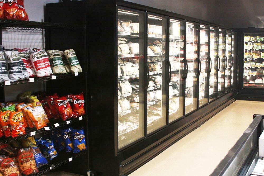 Black industrial shelving unit stocked with snack bags next to a long row of glass-fronted refrigerated freezer doors.