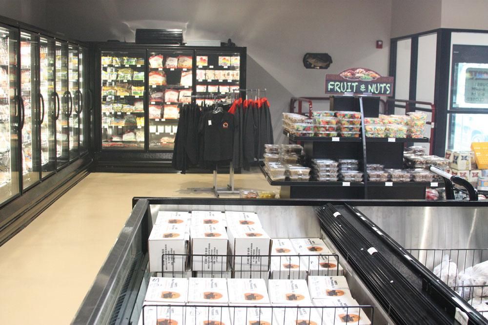 An aisle of a grocery store featuring refrigerated glass doors, apparel on a rack, and a display stand for fruit and nuts.