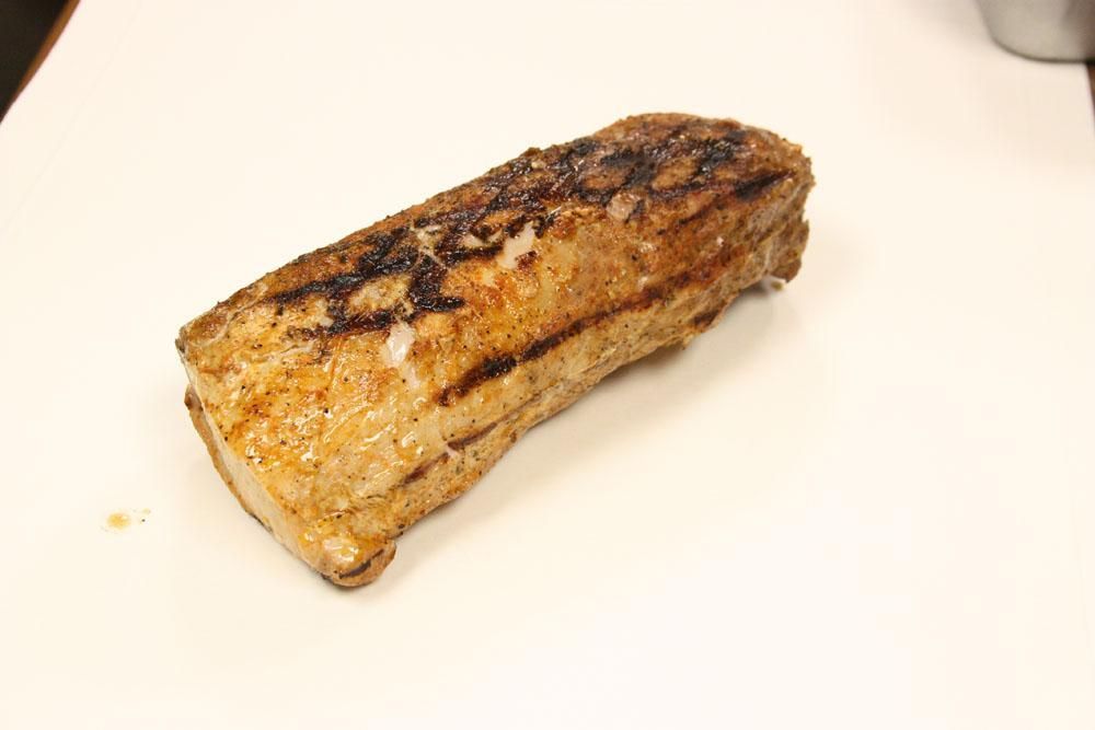 A grilled, rectangular piece of seasoned pork tenderloin sits on a white surface.