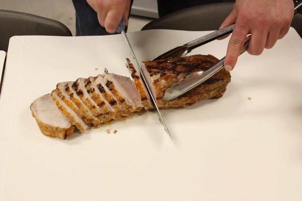 A person slices a grilled pork tenderloin on a white cutting board using a serrated knife and metal tongs.