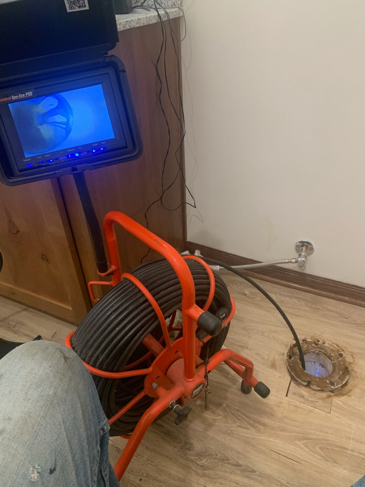 Orange drain snake tool on a floor beside a cabinet and a wall outlet, with a TV screen in the background