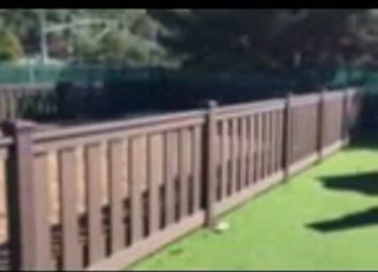 Brown wooden fence along green turf with trees in the background.