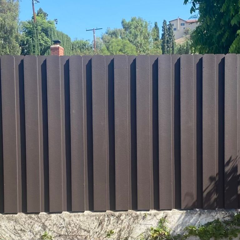 Brown vertical corrugated metal fence in front of a wall, with trees and sky visible in the background.