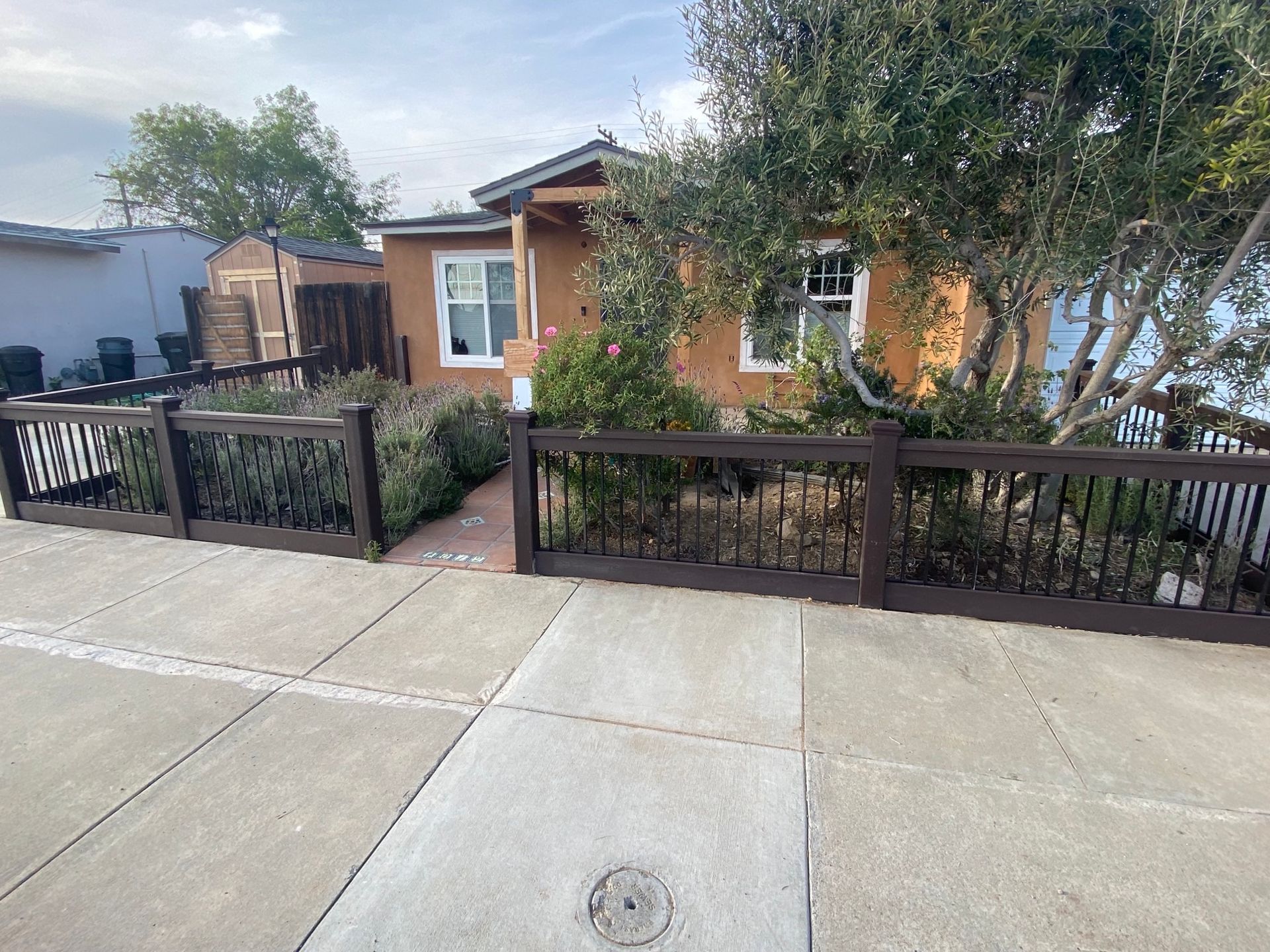 Tan house with brown fence and sidewalk. Tree in the yard.